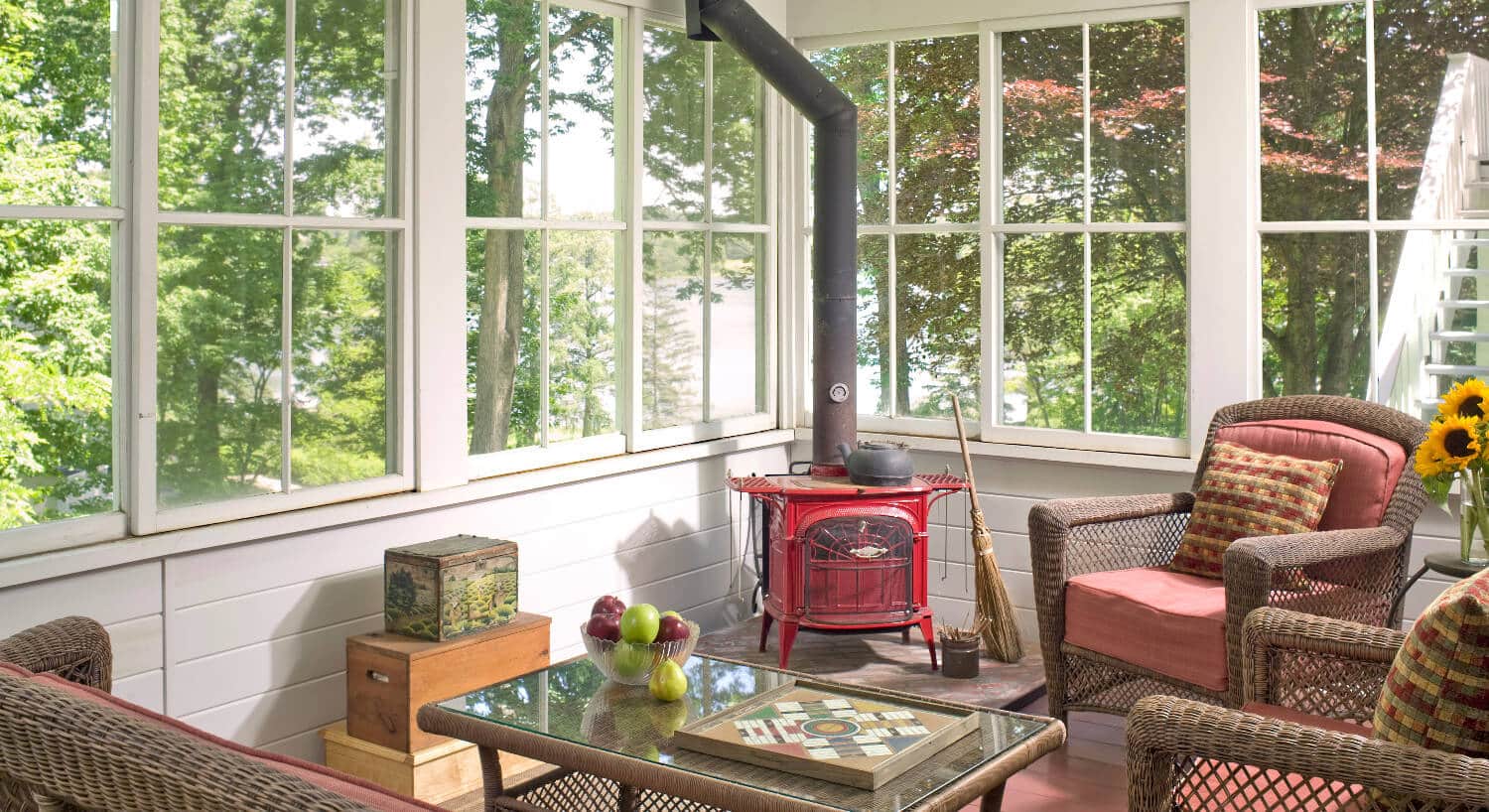 Sunny room with many windows holds cchairs, a table and a red wood stove. 