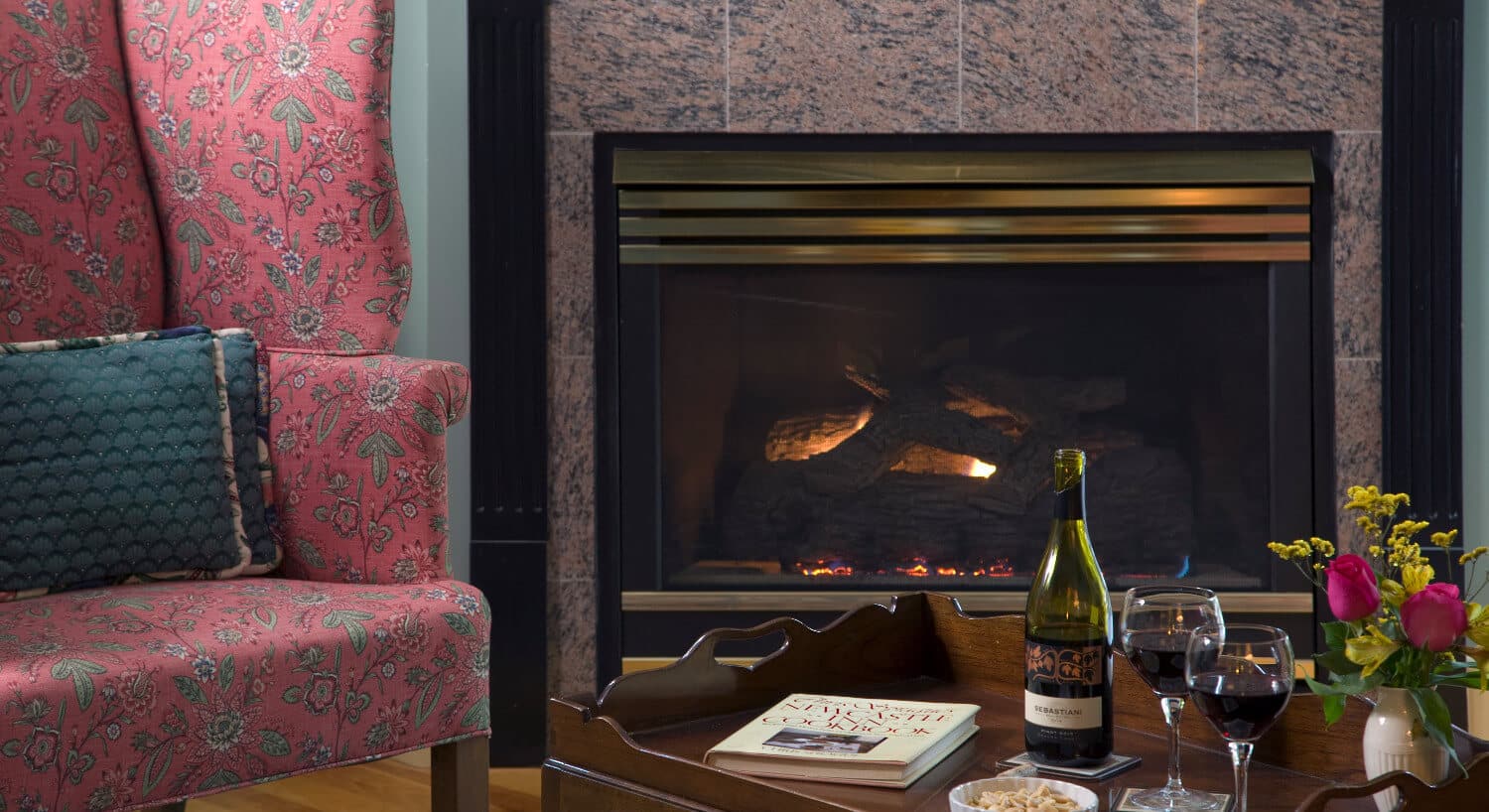 Wingback chairs sits next to a large fireplace fronted by a table with snacks and a bottle of wine. 