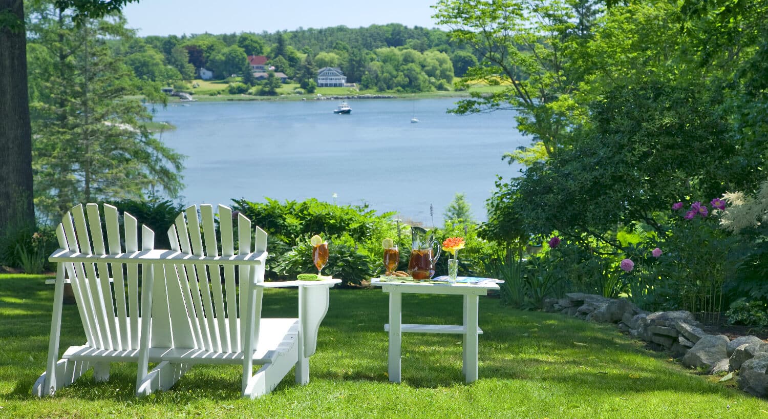 Two white adirondack chairs next to a small table on the grass overlooking the water.
