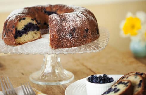 Blueberry bundt cake on a glass dish with a slice of cake and bowl of berries on a plate alongside.