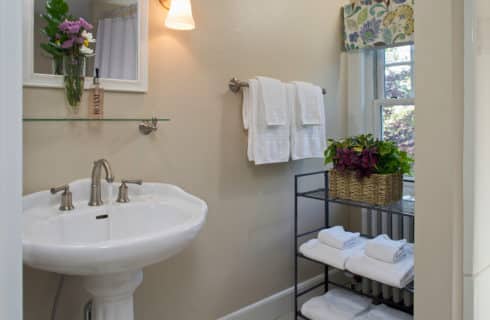 Bathroomw ith a white pedestal sink and mirror and a cart holding a plant and extra towels.