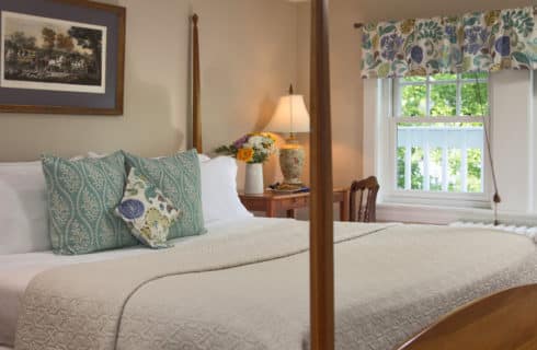 Large four-post bed made up in cream with decorative pillows next to a table and desk beside a large window.