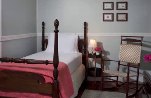 Cozy bedroom with wooden four-post bed made up in white and coral bedding with a dark wood side table and rocking chair.