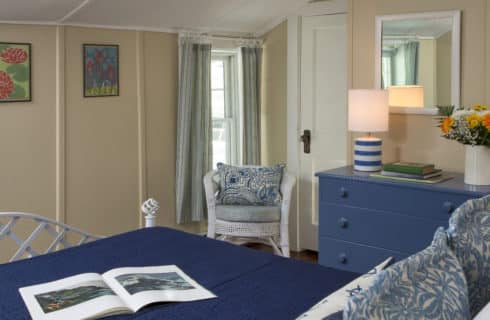 Cozy warm bedroom with a white iron bed made up in navy blue with a blur dresser and white wicker chair.