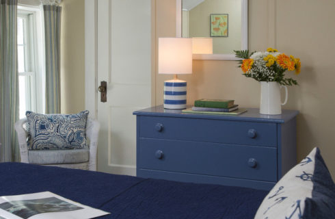 Cozy warm bedroom with a white iron bed made up in navy blue with a blur dresser and white wicker chair.