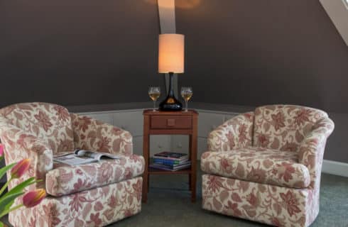 Seating area with two pattterned over-stuffed chairs and a table with a lamp and wine glasses.