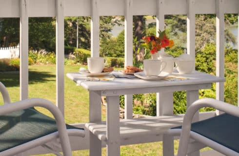 Deck with white rails holds a small white wooden table and chairs set with coffee cups and flowers.