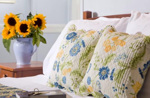 Bed with blue yellow and white shams alongside a bedside table with a vase full of sunflowers.