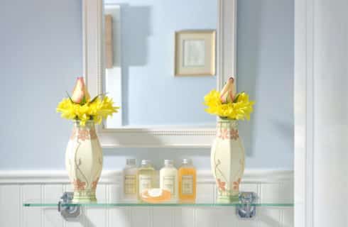 Glass shelf in bathroom with elegant vases full of yellow flowers and a white-framed mirror.