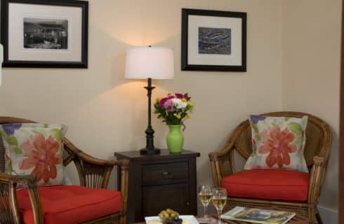 Wicker chairs with red cushions form a seating area with a small table n the corner of a bedroom.