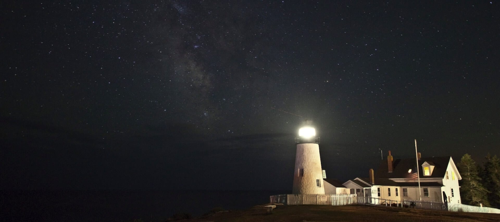 A lighthouse is lit up in the dark of night, surrounded by stars and the Milky Way.