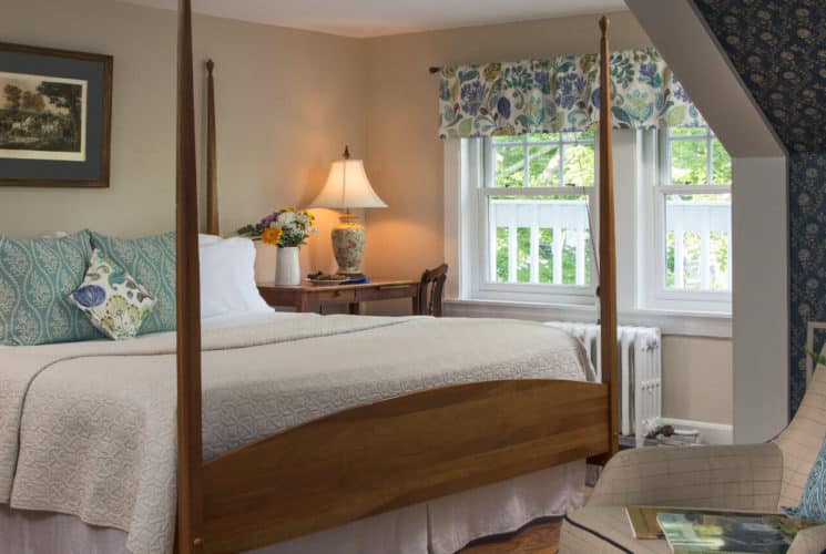 Large four post wooden bed in a beige and blue bedroom with a large window and pitched roof.