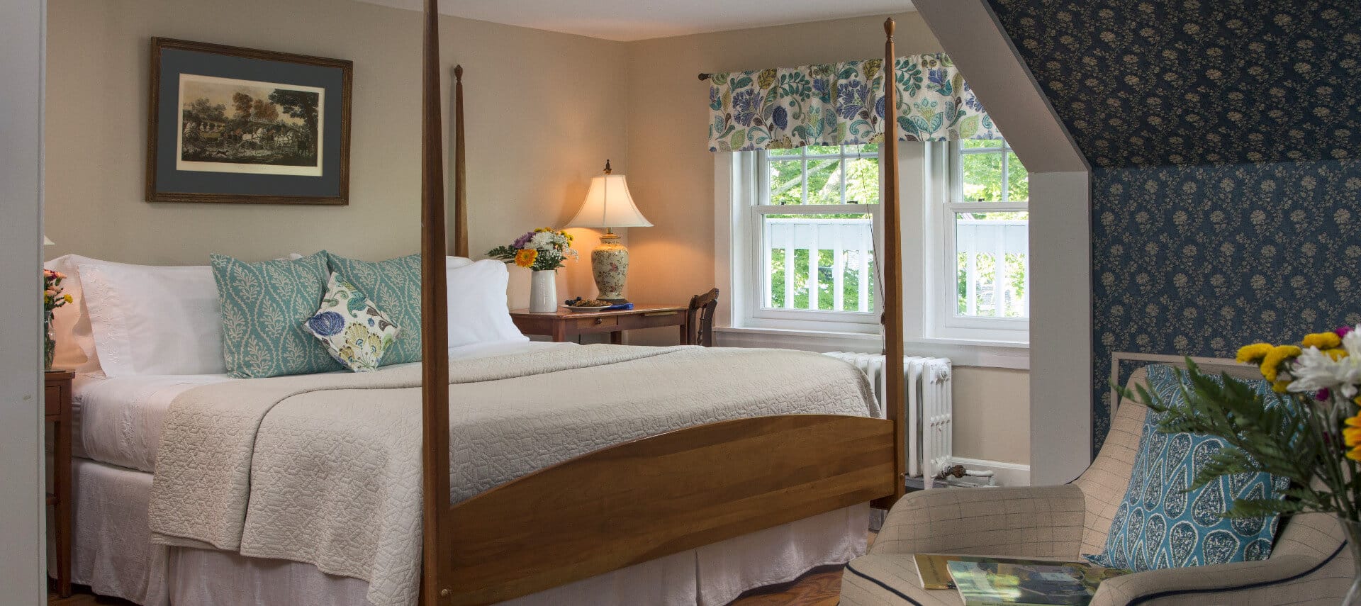 Large four post wooden bed in a beige and blue bedroom with a large window and pitched roof.