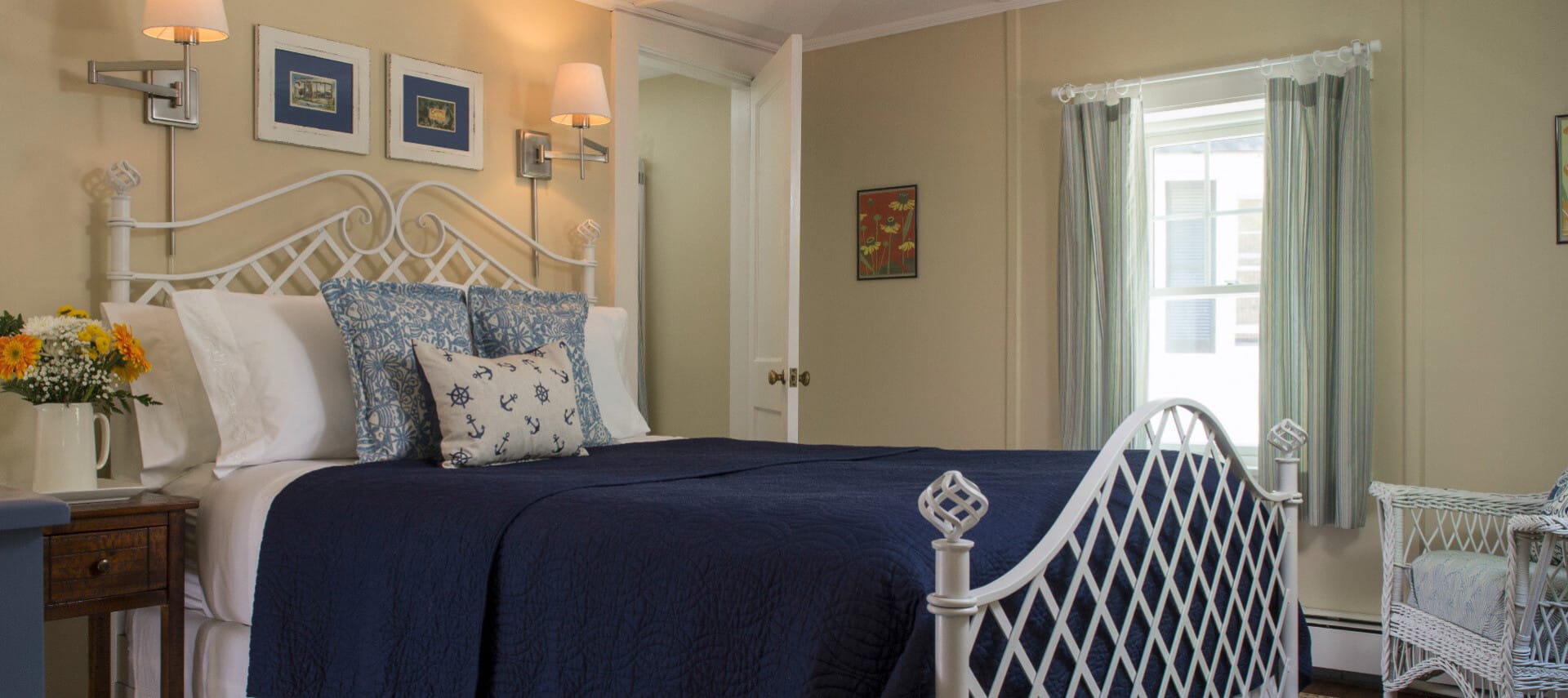 Cozy warm bedroom with a white iron bed made up in navy blue with a blur dresser and white wicker chair.