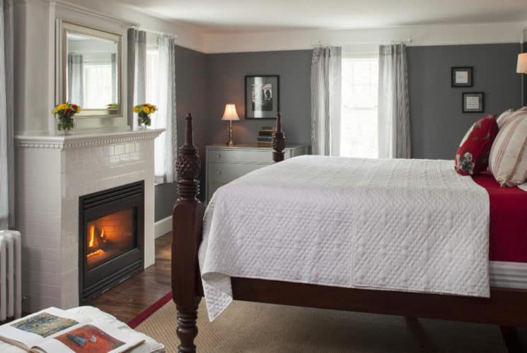 Large four-post bed made up in white in a room with grey walls and white crown molding and a white fireplace..