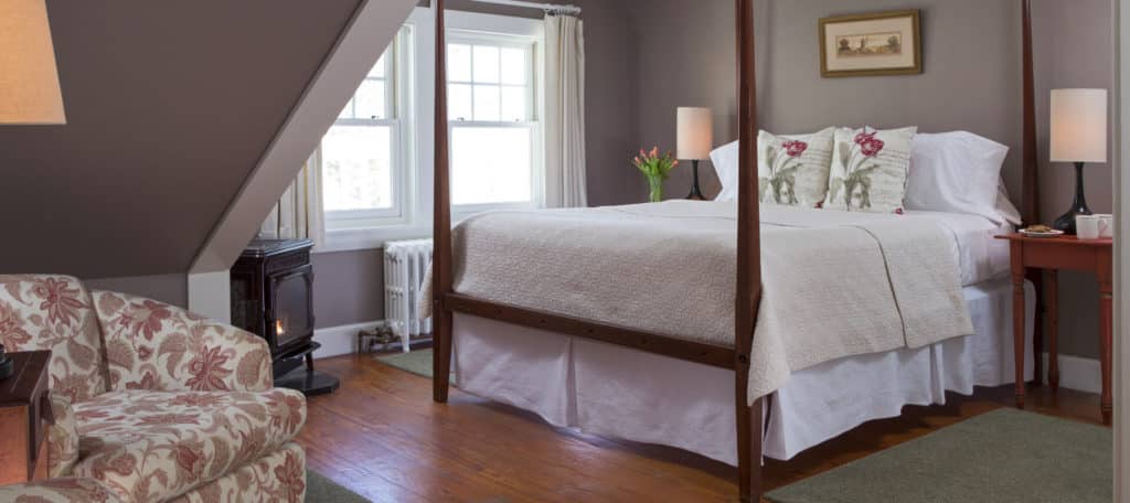 Gracious room painted in grey holds a large four-post bed with white bedding, and a small seating area under a pitched roof.