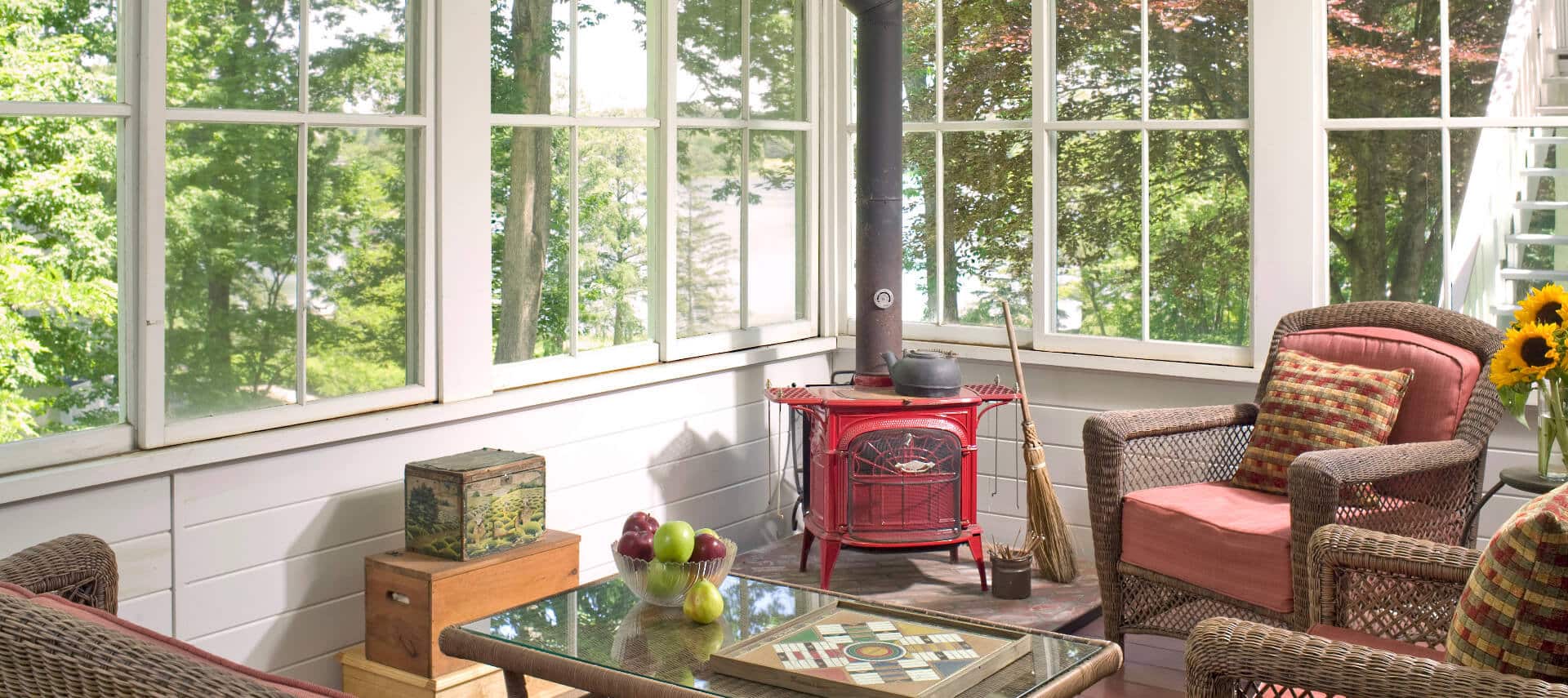 Sunny room with many windows holds cchairs, a table and a red wood stove.
