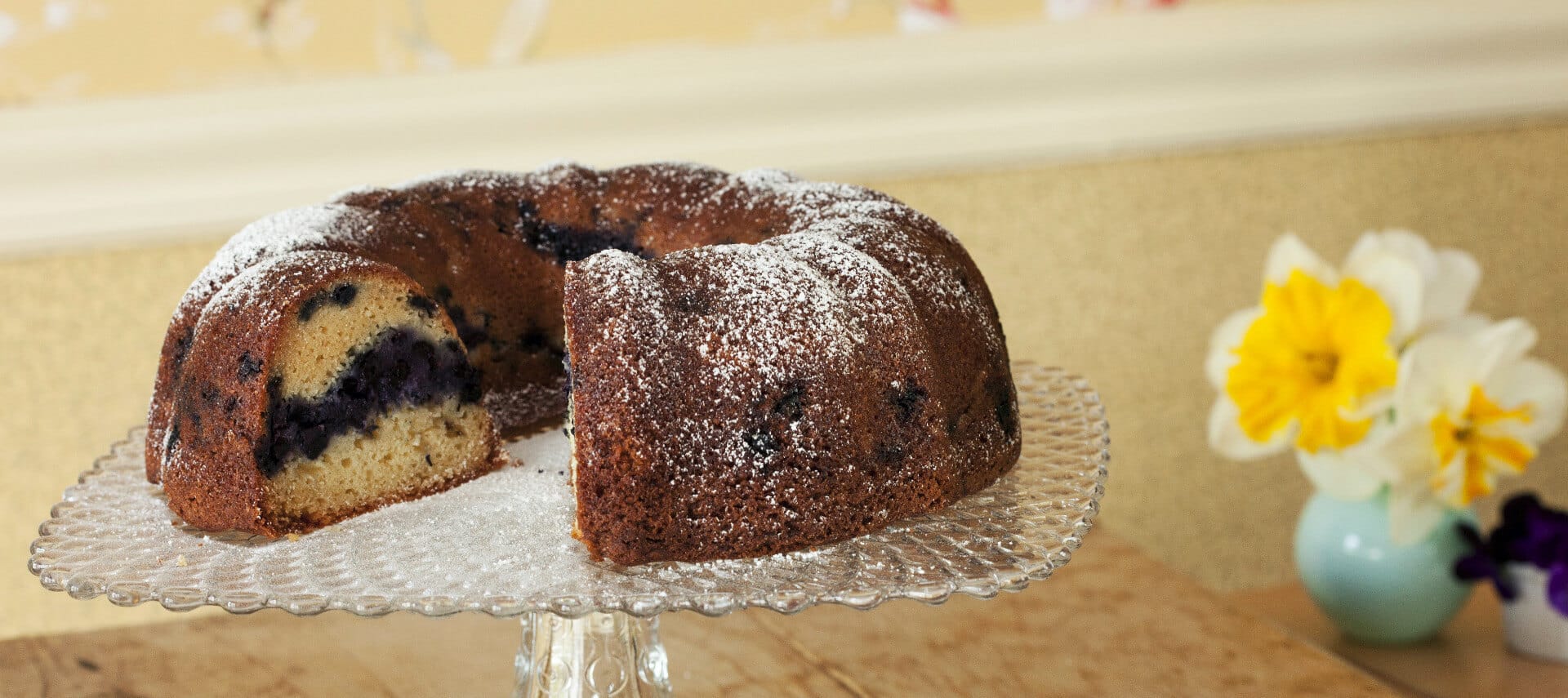 Blueberry bundt cake on a glass dish with a slice of cake and bowl of berries on a plate alongside.