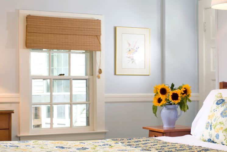 Bright cheerful bedroom with wooden bed made up in white yellow and blue quilt and shams with a large window and wooden dresser.