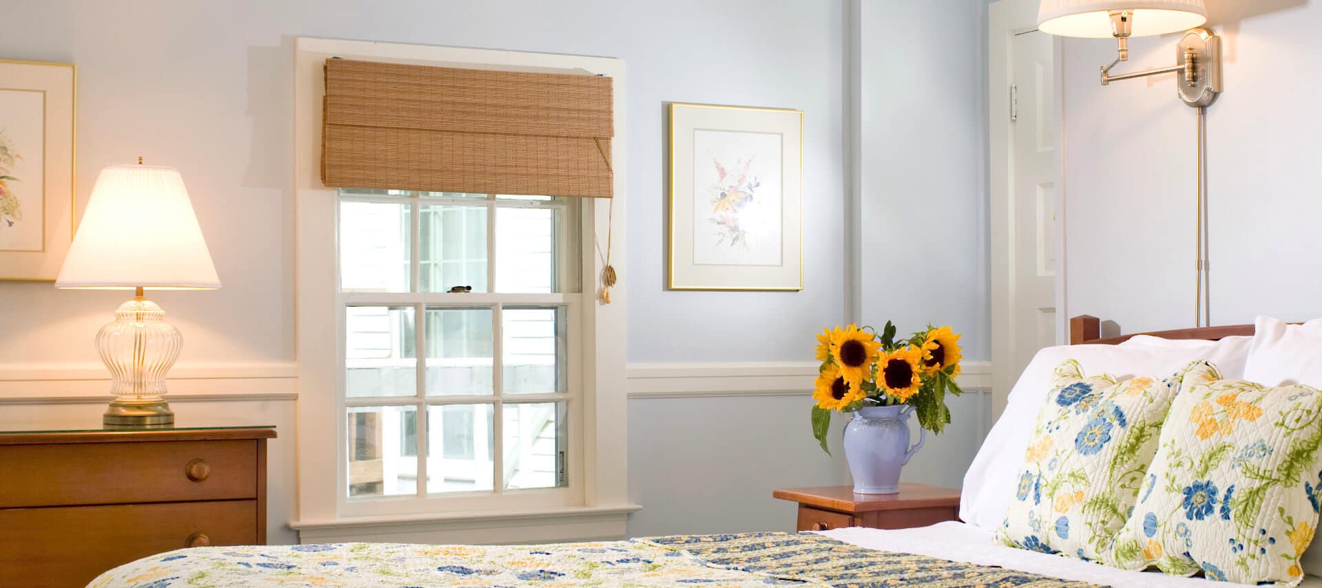 Bright cheerful bedroom with wooden bed made up in white yellow and blue quilt and shams with a large window and wooden dresser.