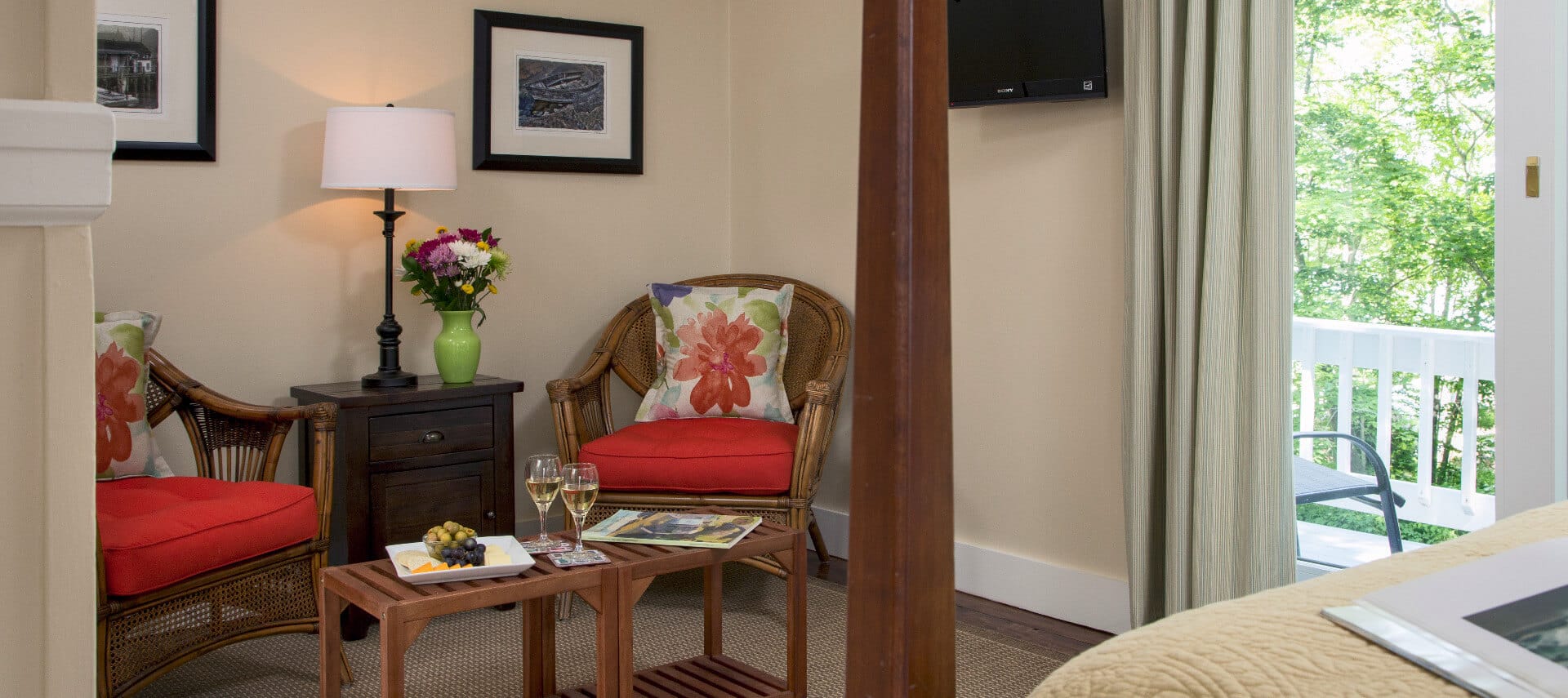 Wicker chairs with red cushions form a seating area with a small table n the corner of a bedroom.