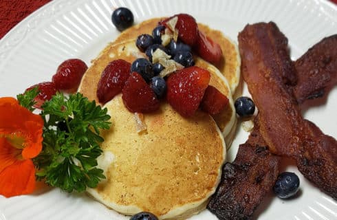 White plate on red tablecloth with three fluffy pancakes topped with strawberries and blueberries and butter dollop next to thick sliced bacon.