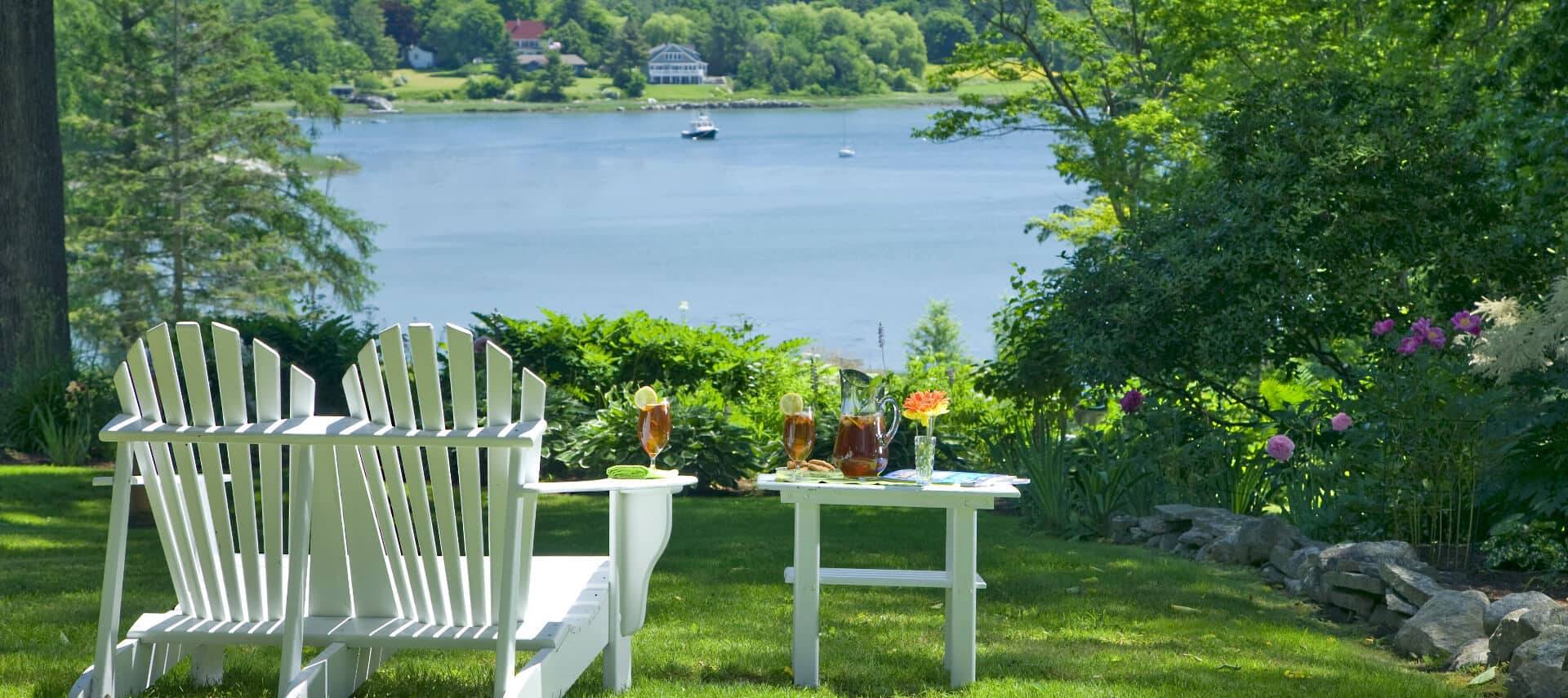 Two white adirondack chairs next to a small table on the grass overlooking the water.