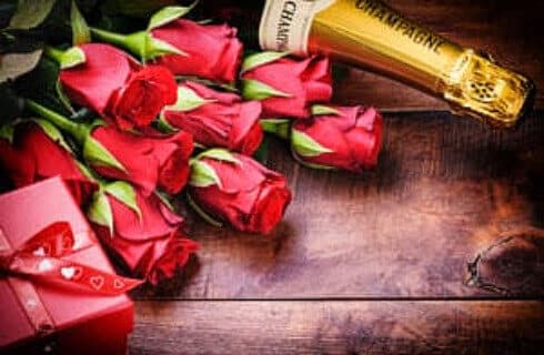 Red roses on a wooden table with a bottle of champagne and a gift wrapped in red.